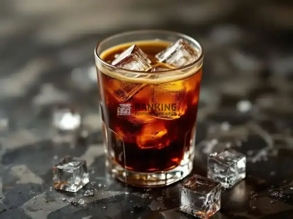 Szklanka tonic espresso z kostkami lodu na ciemnym tle. Napój ma bursztynowy kolor, a wokół znajdują się rozsypane kostki lodu.